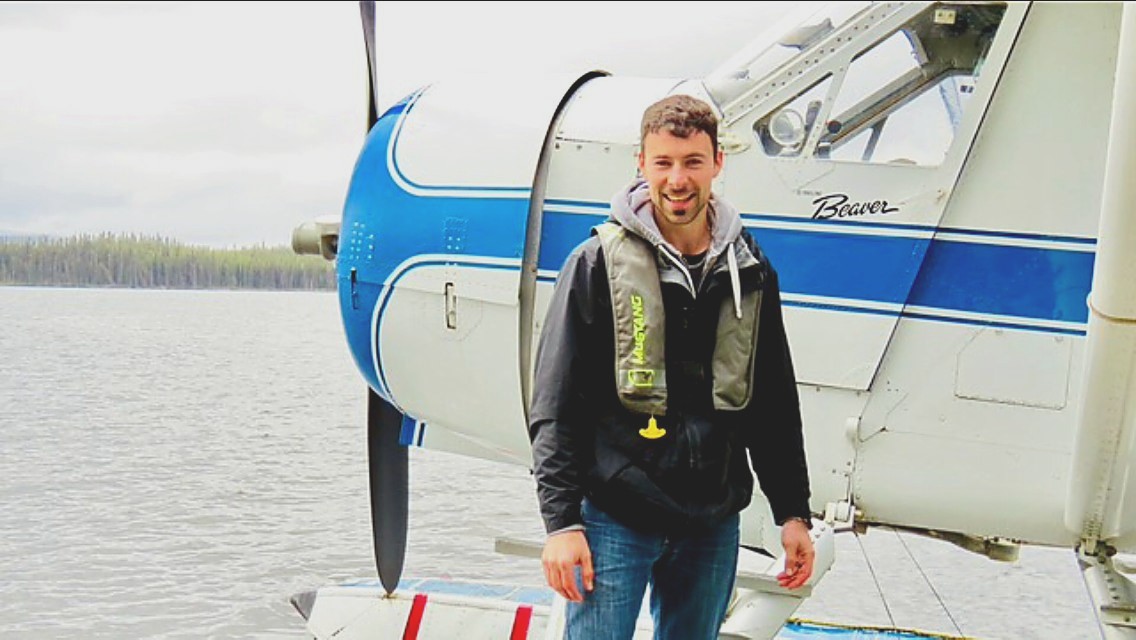 Joe Noble standing on the float of a blue and white de Havilland Beaver at a lake.
