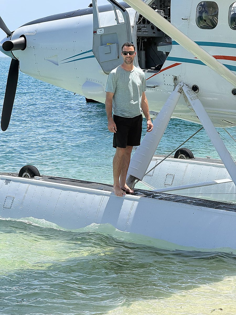 Joe Noble standing on the float of a white seaplane in shallow water.